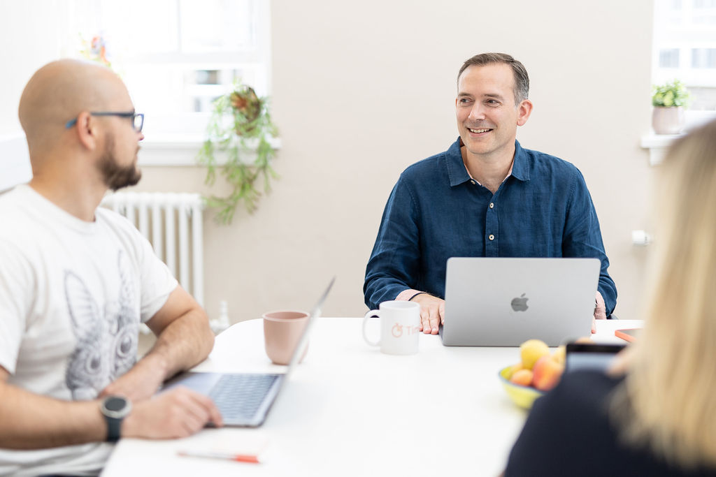 A woman stands smiling next to a man engaged into working on a laptop at a white windowsill in a bright, modern office.